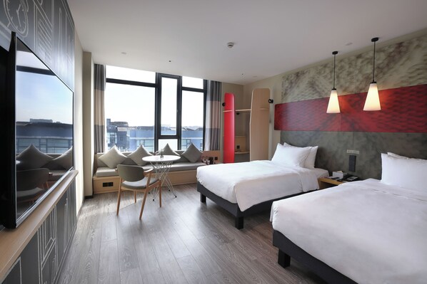 Habitación de hotel con dos camas, zona de estar con una mesa, un ventanal y vistas a la ciudad.