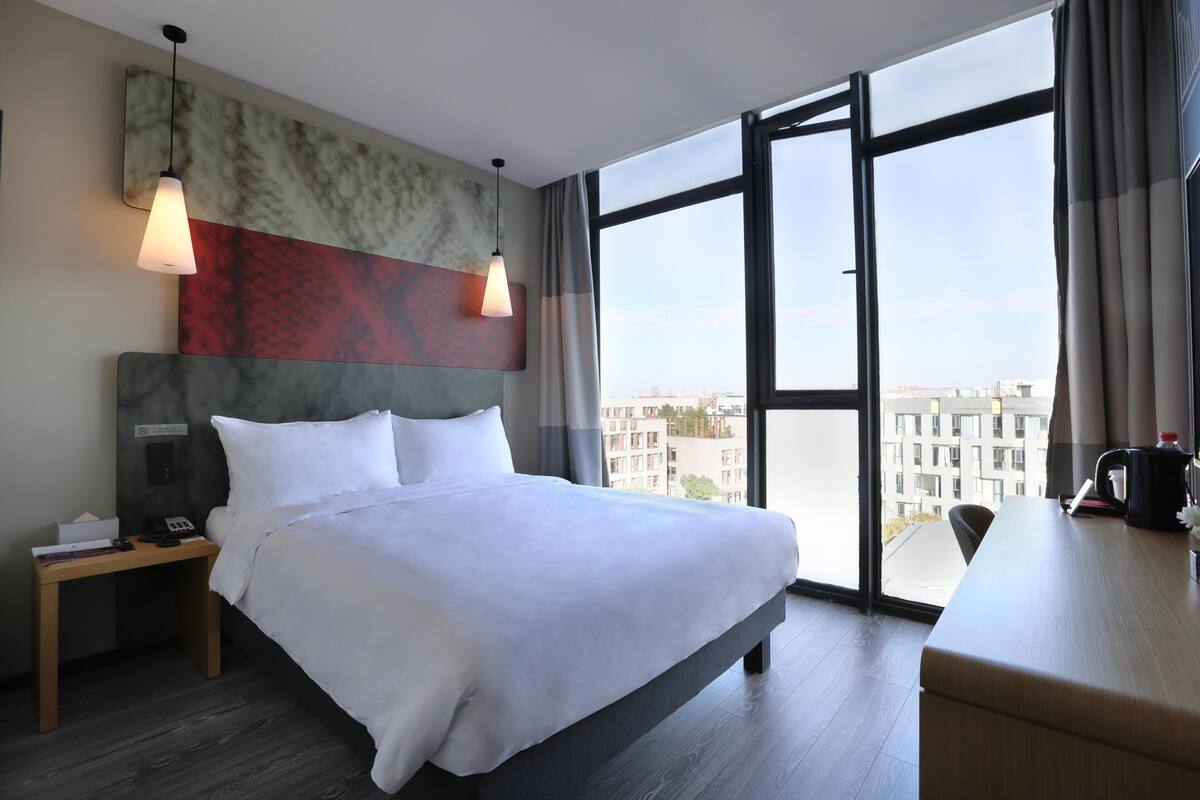 A hotel room with a bed, bedside table, desk, and large windows offering a city view.