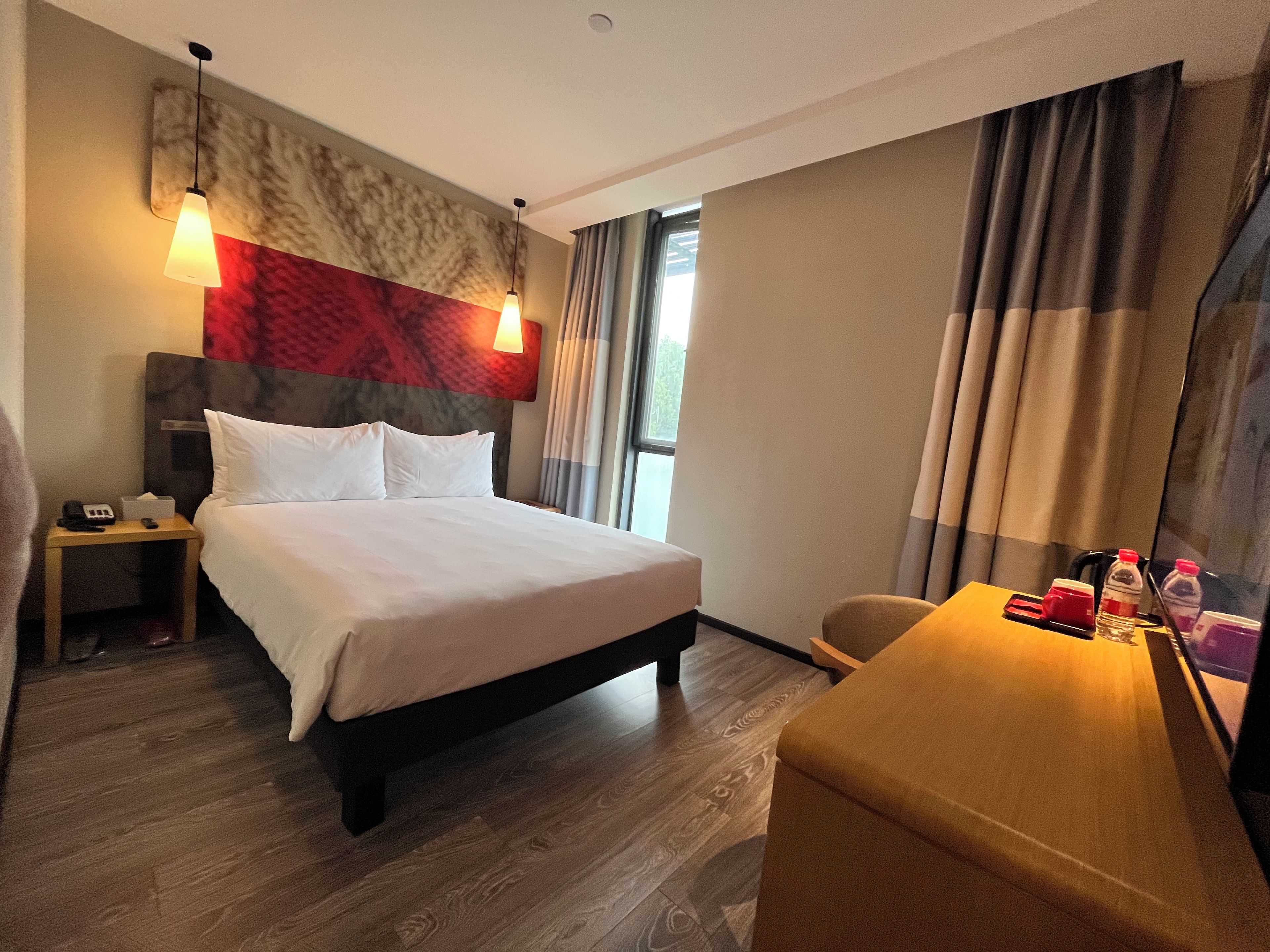 A hotel room with a bed, bedside table, lamp, and a desk with a chair.