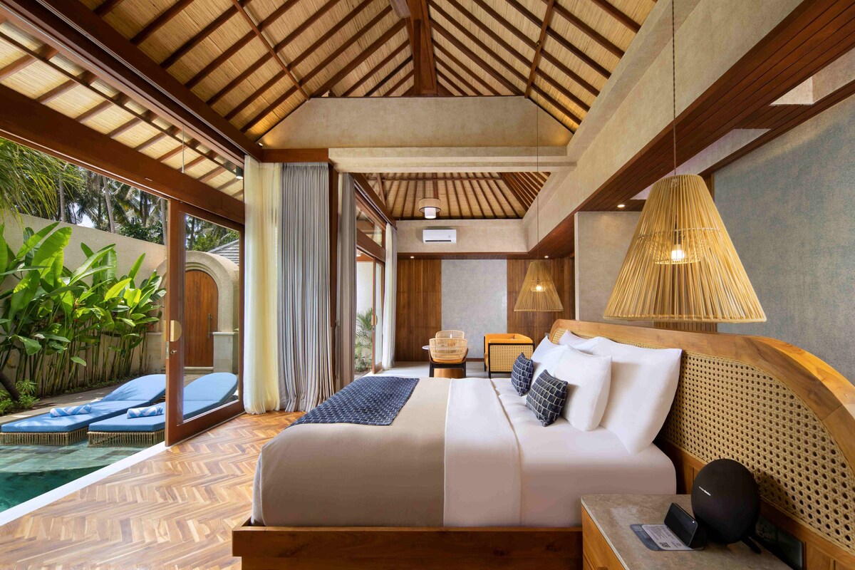 A bedroom with a bed, bedside table, lamp, and a view of an outdoor area with a pool.