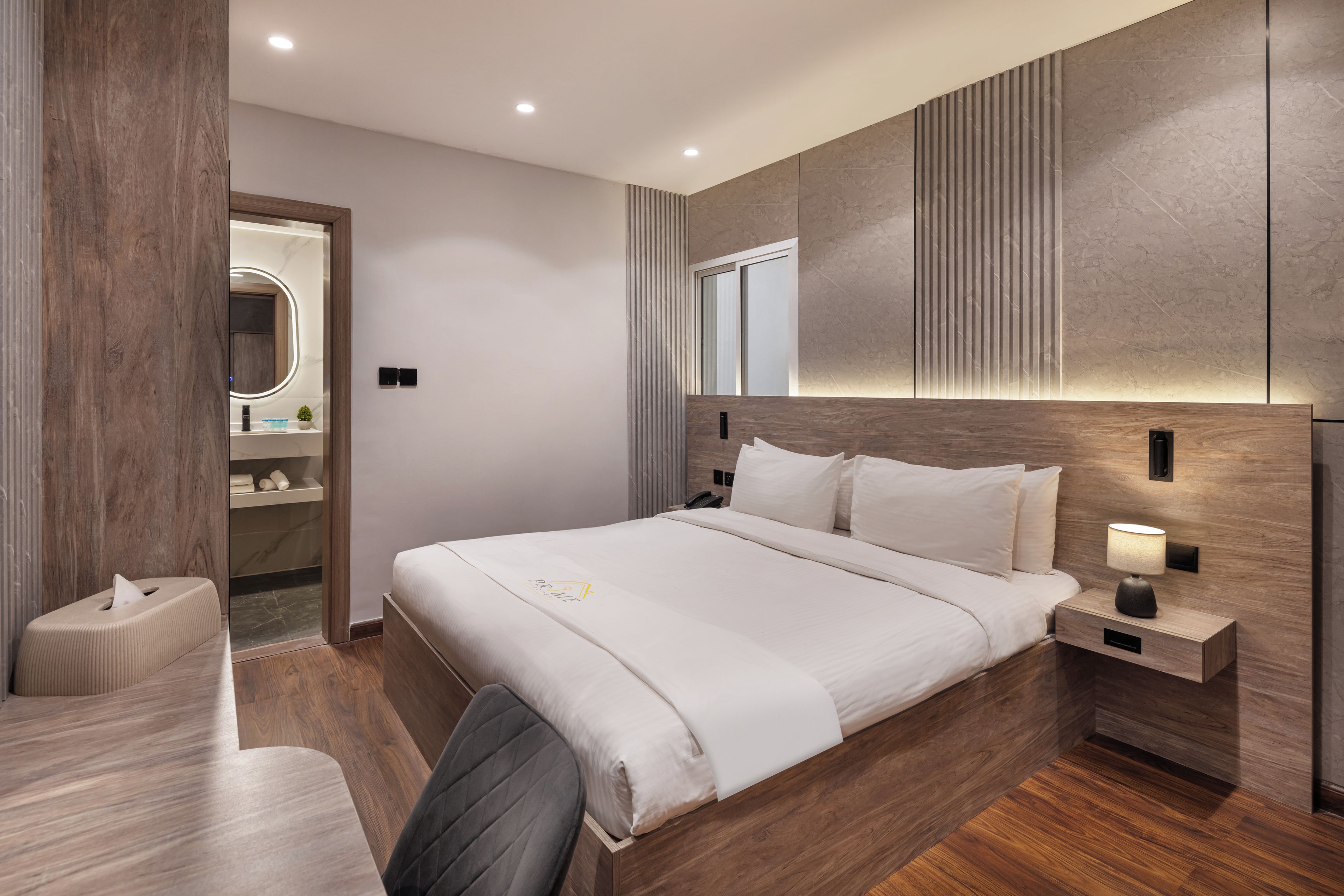 Una habitación de hotel moderna con una cama grande, mesita de noche con lámpara, un escritorio pequeño y una silla.