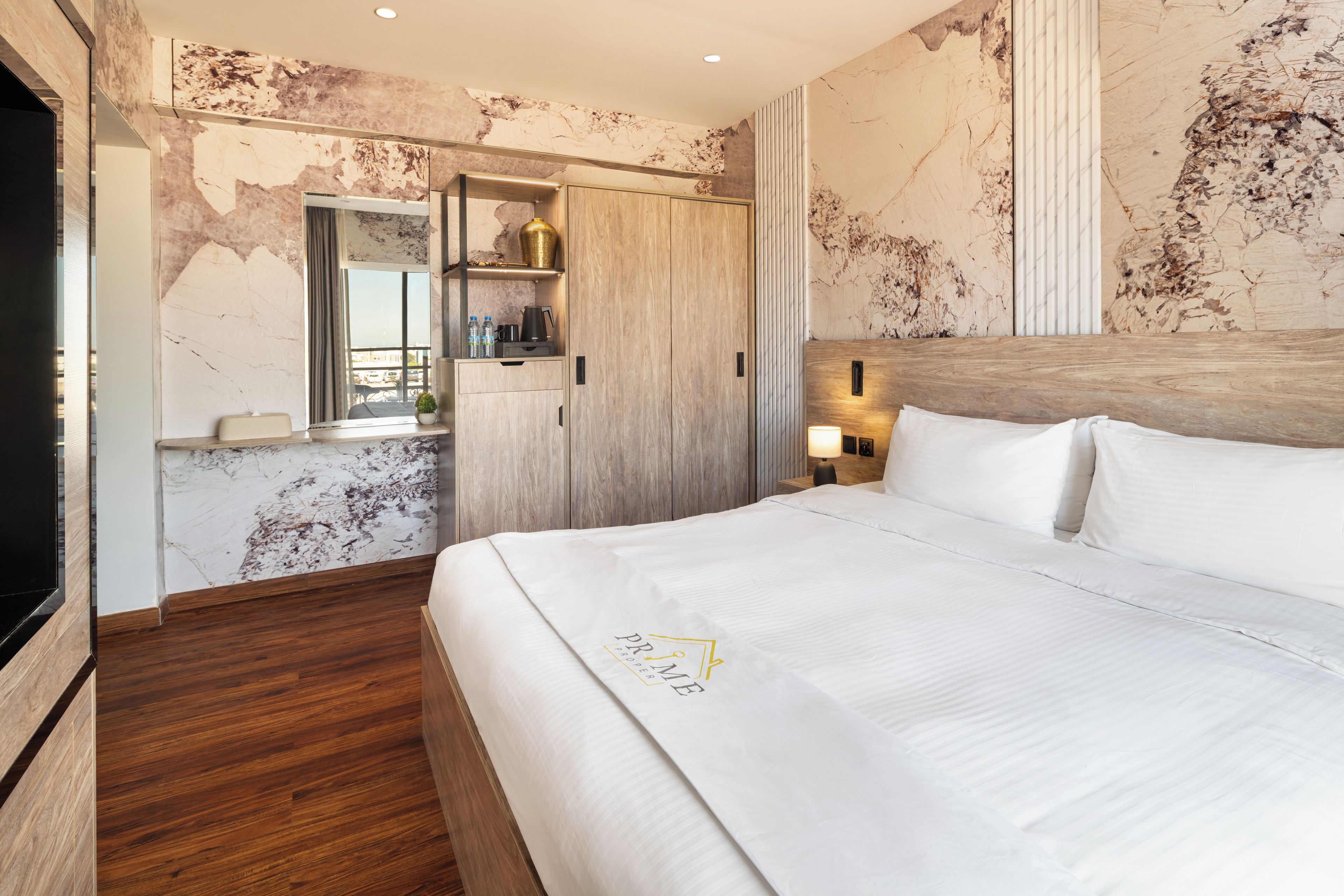 A modern hotel room with a large bed, a wooden wardrobe, a vanity with a mirror, and a marble wall.