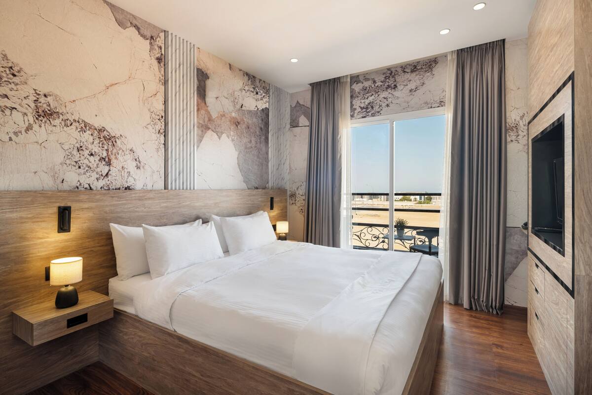 Habitación de hotel con una cama grande, televisión, mesita de noche con lámpara y vista al exterior.