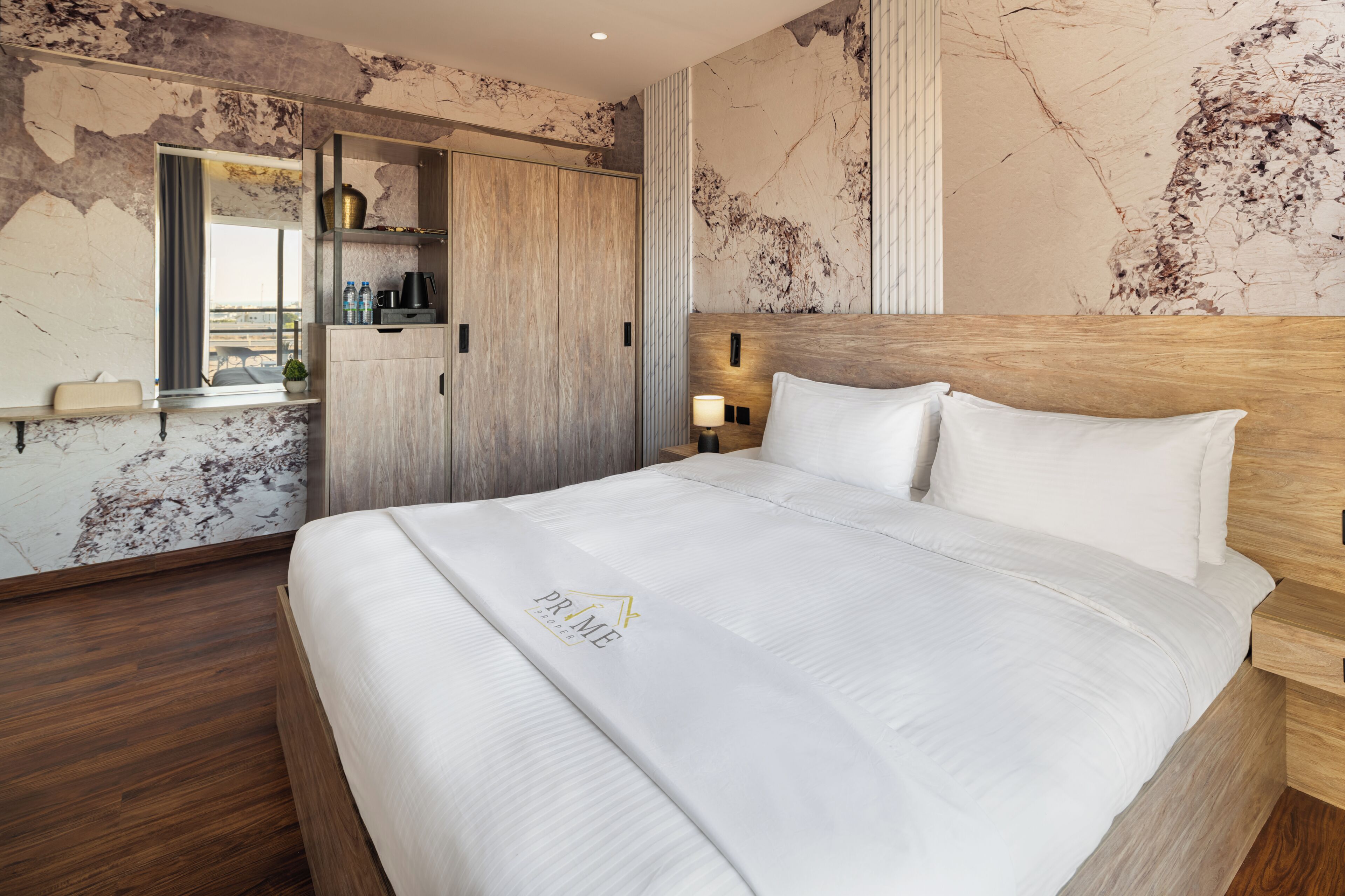 A hotel room with a large bed, a wooden headboard, a built-in wardrobe, a bedside table with a lamp, and a marble wall.