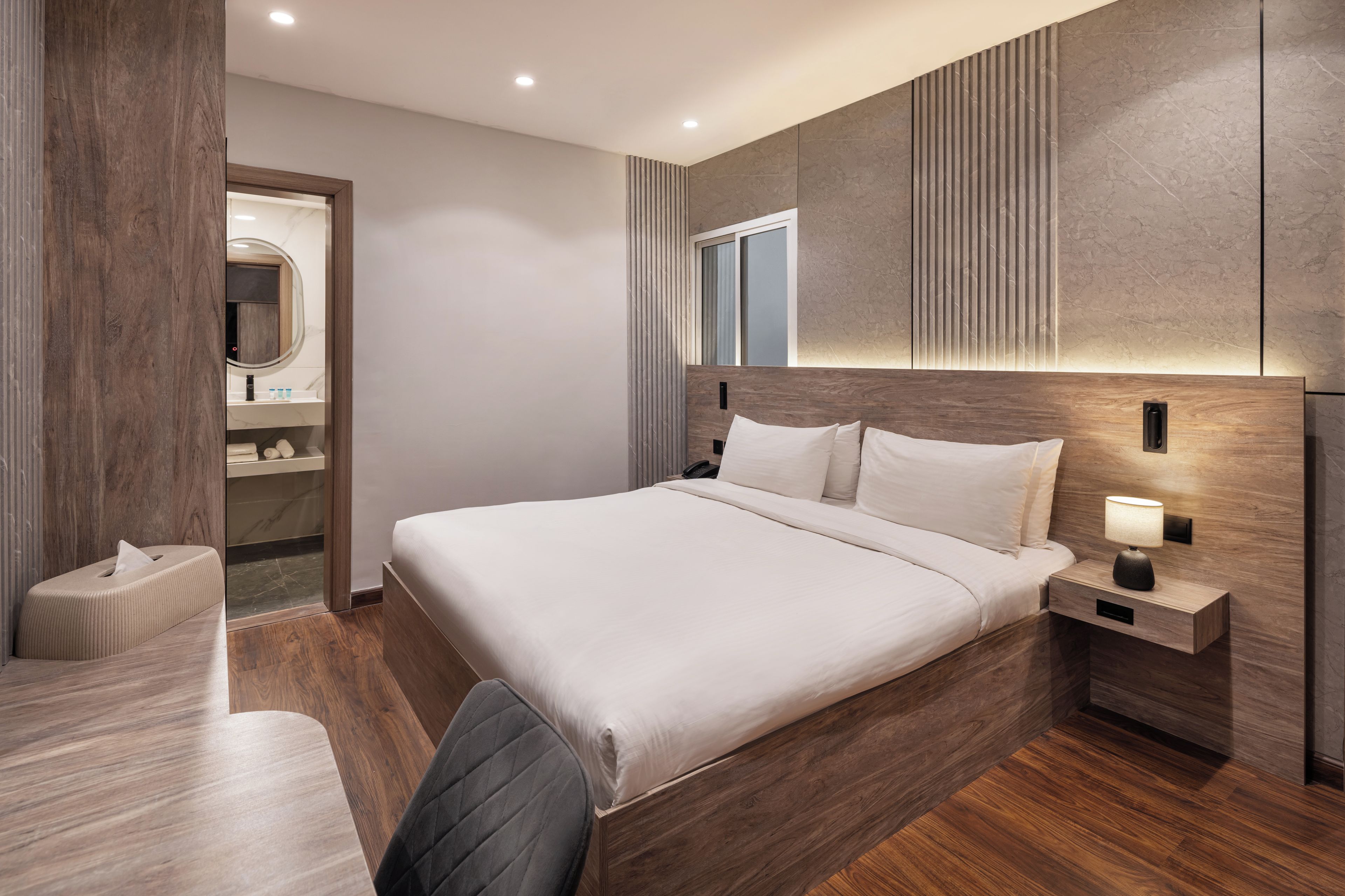 A modern hotel room with a large bed, bedside table with a lamp, a small bedside table, a chair, and a bathroom visible through an open door.