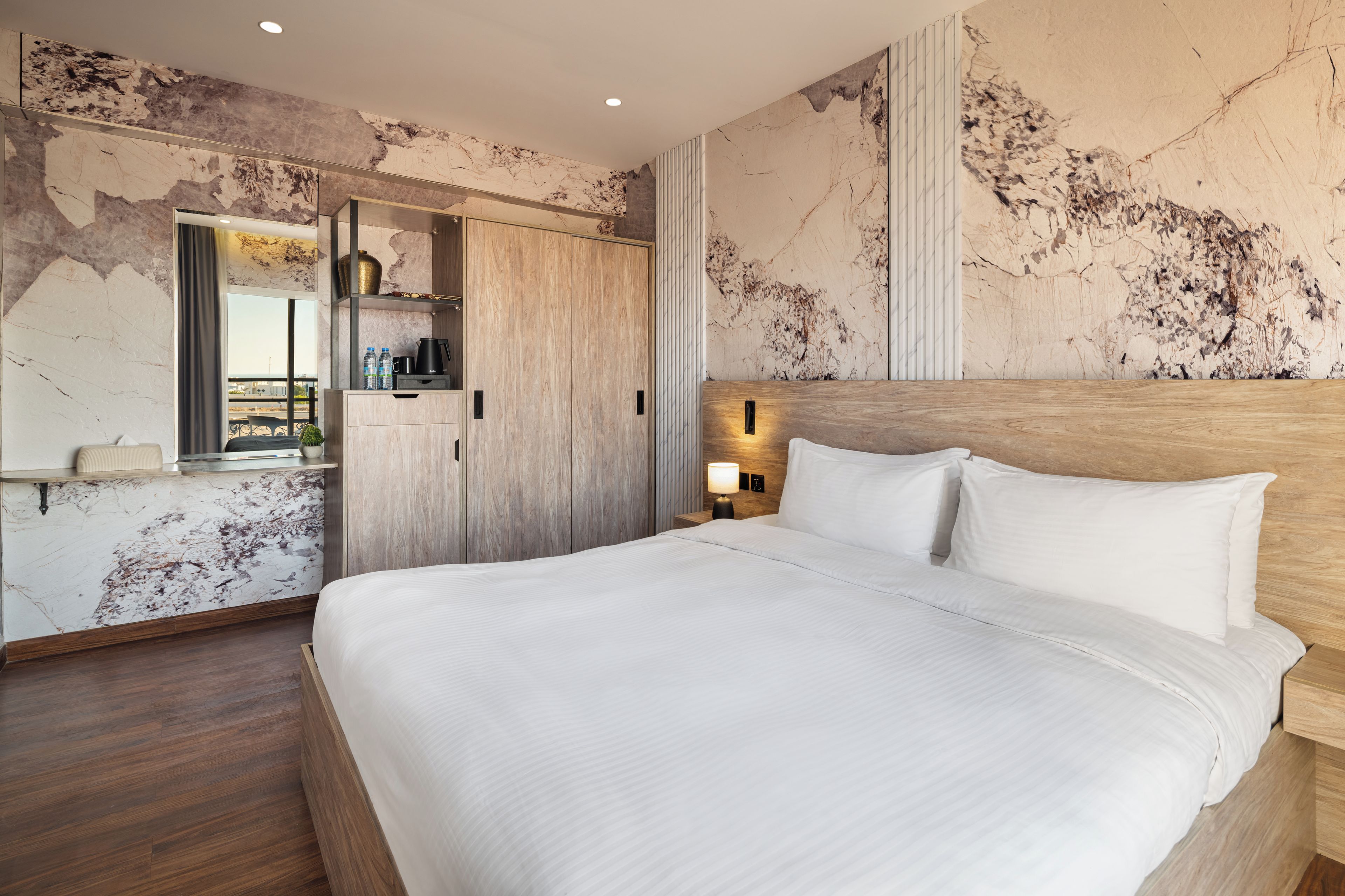 A modern hotel room with a large bed, a wooden headboard, a nightstand with a lamp, a wardrobe, and a marble wall.