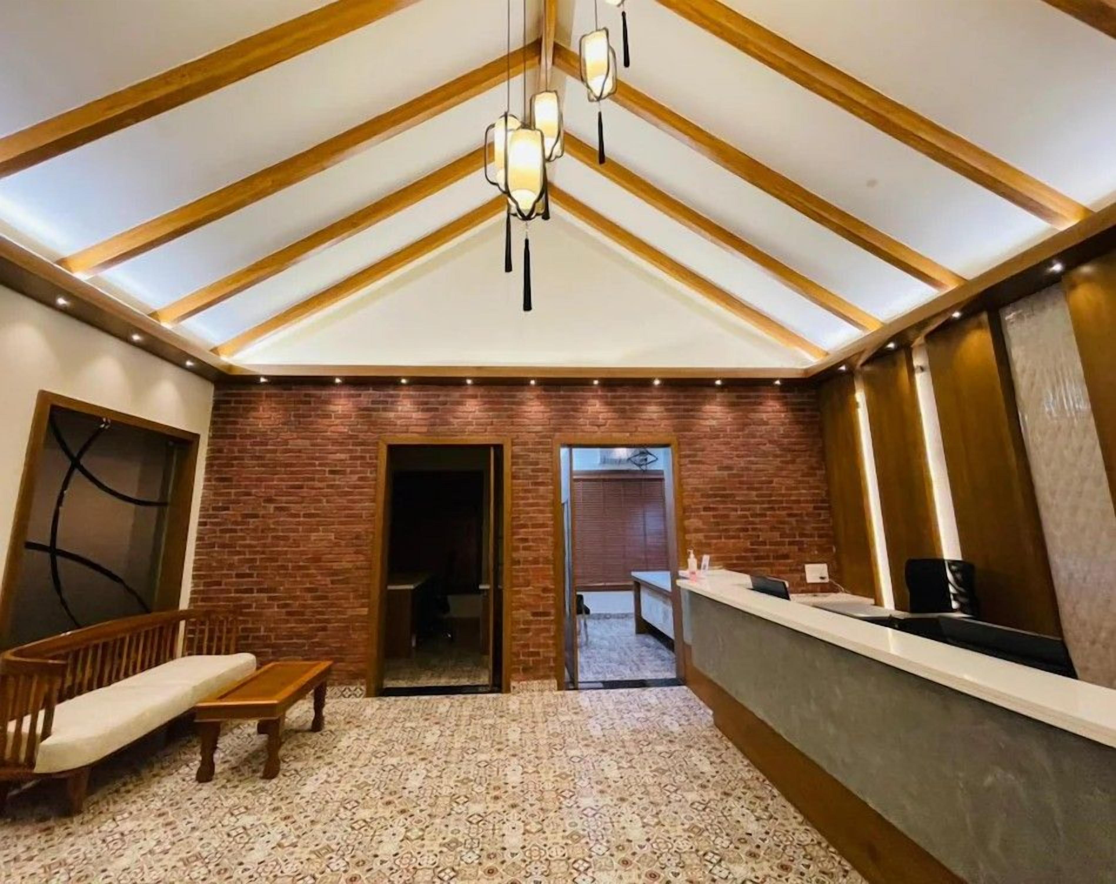 A room with a high ceiling, exposed wooden beams, a brick wall, a reception desk, a bench, and a patterned carpet.