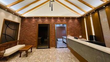 A room with a high ceiling, exposed wooden beams, a brick wall, a reception desk, a bench, and a patterned carpet.