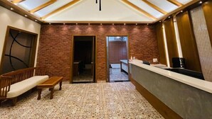 A room with a high ceiling, exposed wooden beams, a brick wall, a reception desk, a bench, and a patterned carpet. - Aara Jungle Resort (Mettupalayam)