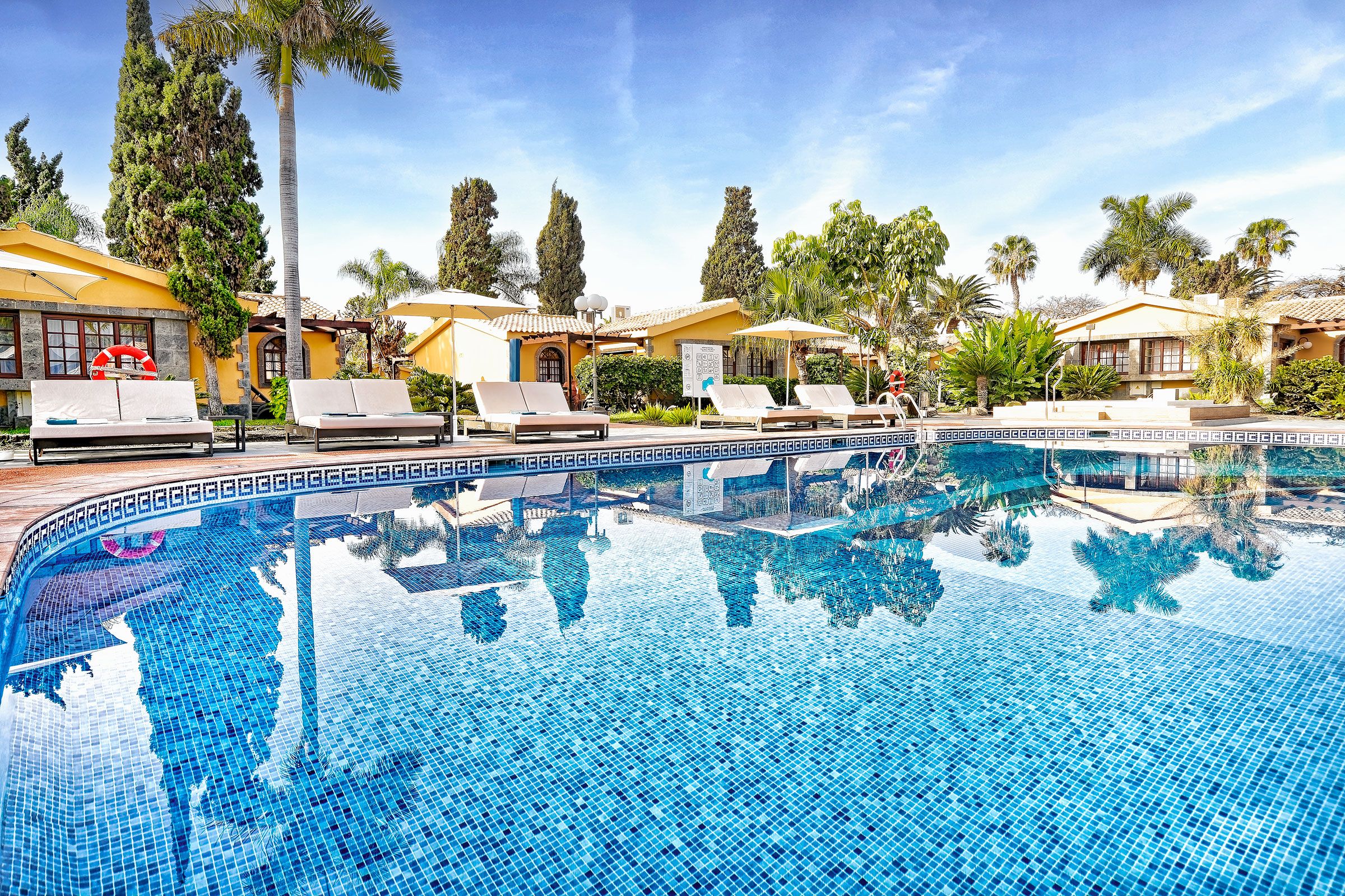 A swimming pool with lounge chairs and umbrellas, surrounded by palm trees and a clear sky.