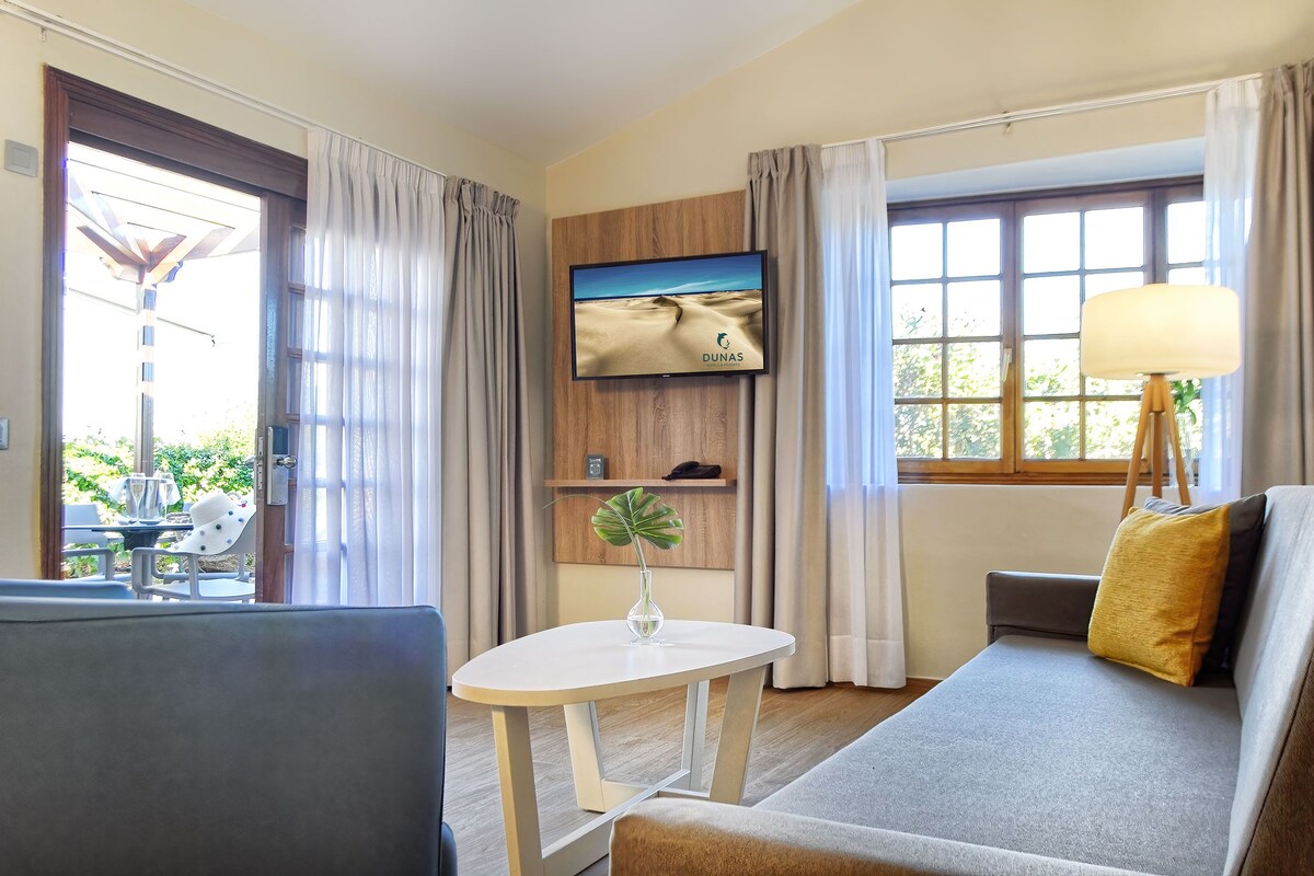 Quarto de hotel com sofá, uma pequena mesa com um vaso, televisão na parede e janela com cortinas.