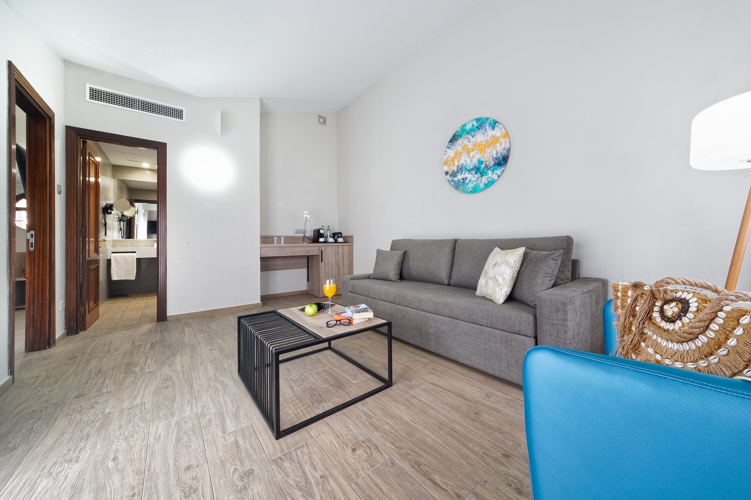 Un salón con un sofá gris, un sillón azul, una mesa de centro y una cocina al fondo.