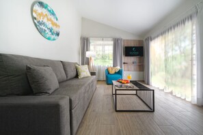 Ein modernes Wohnzimmer mit einem grauen Sofa, einem blauen Sessel, einem Couchtisch mit einer Fruchtplatte und einem Glas Orangensaft, einem Fernseher an der Wand und einem großen Fenster mit Vorhängen.