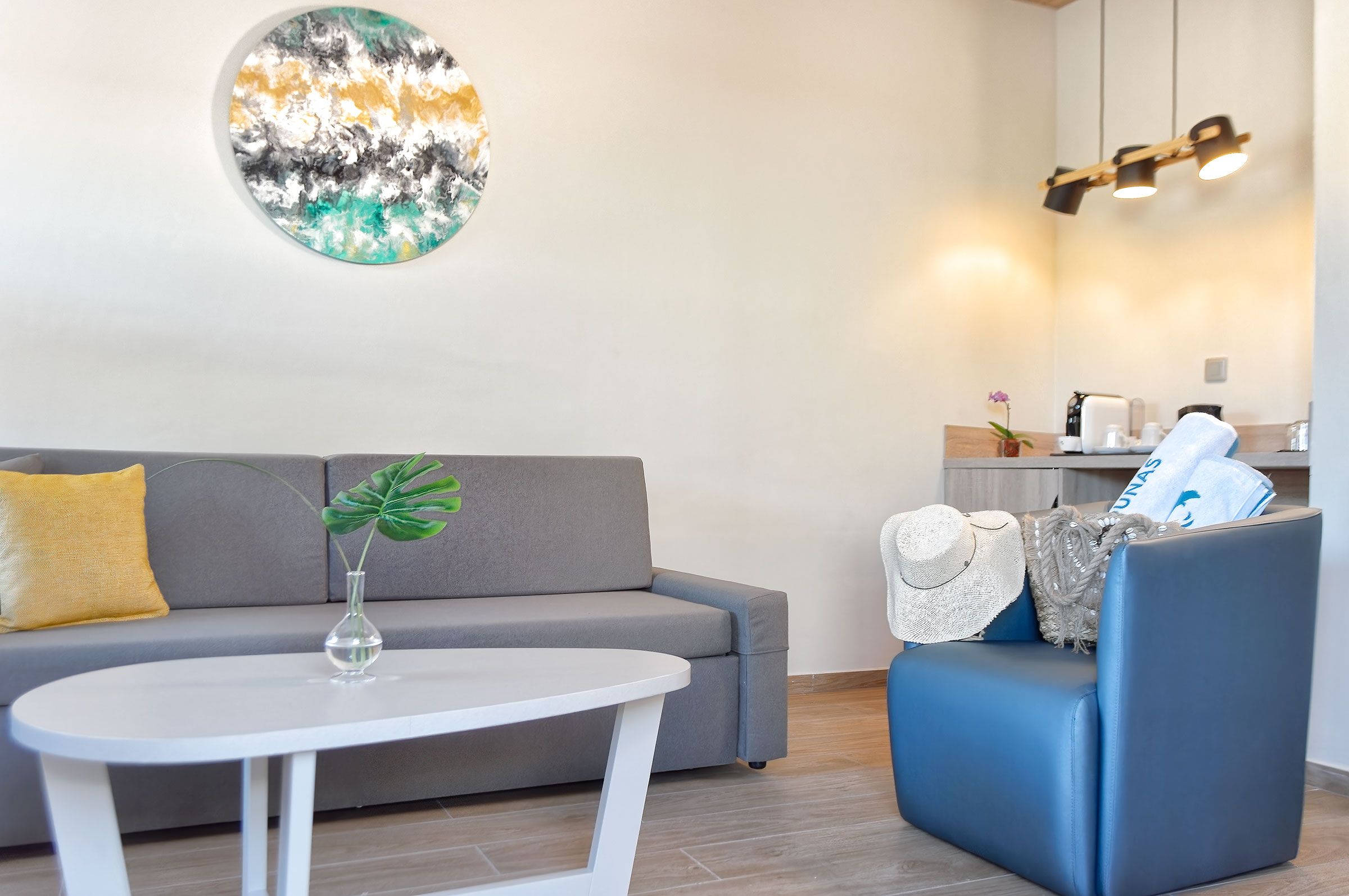 Una sala de estar moderna con un sofá gris, una mesa de centro blanca y un sillón azul. Un cuadro abstracto circular cuelga en la pared, y se distingue un pequeño rincón de cocina al fondo.