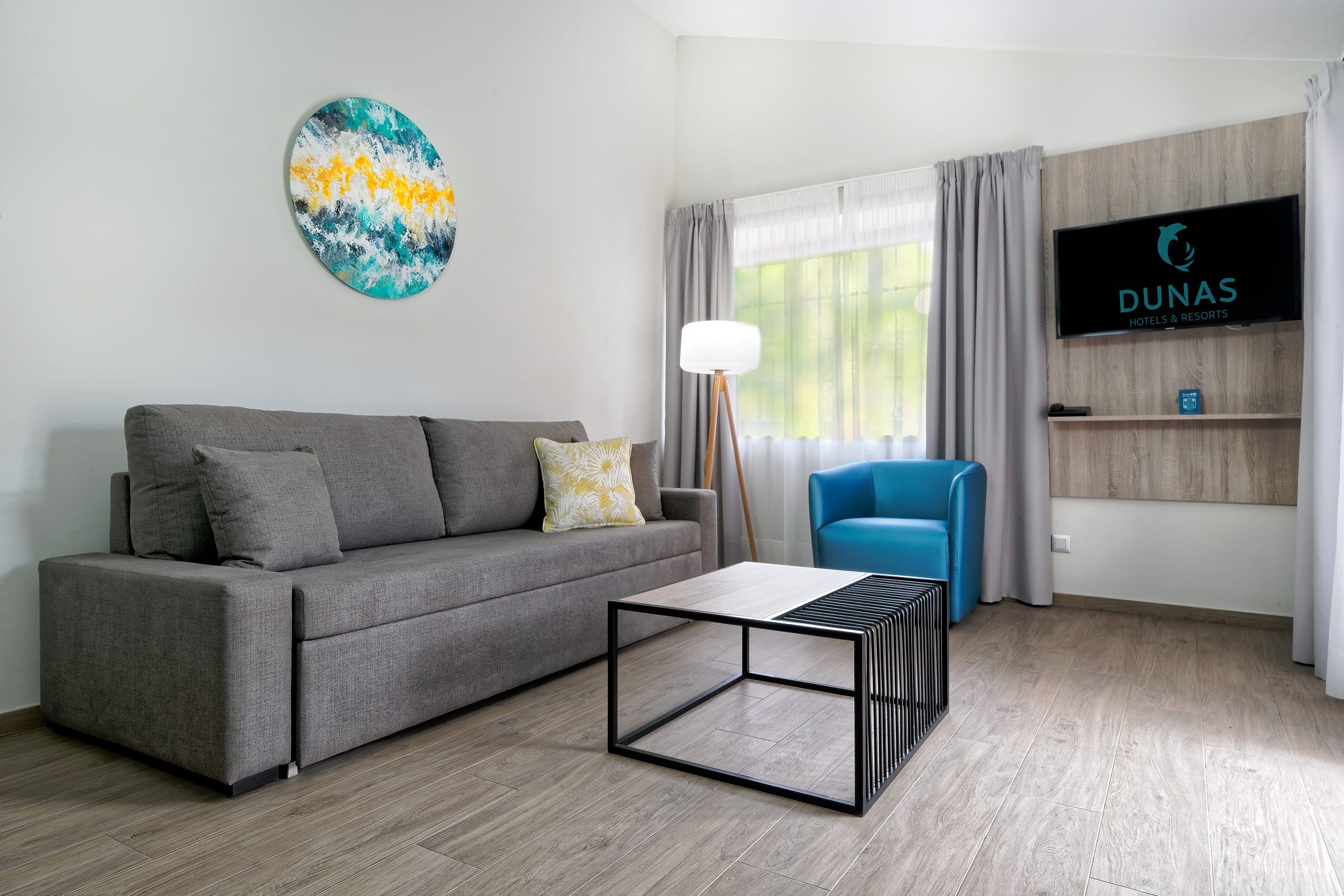 Un salon moderne avec un canapé gris, une table basse, un fauteuil bleu, un téléviseur et un tableau accroché au mur.
