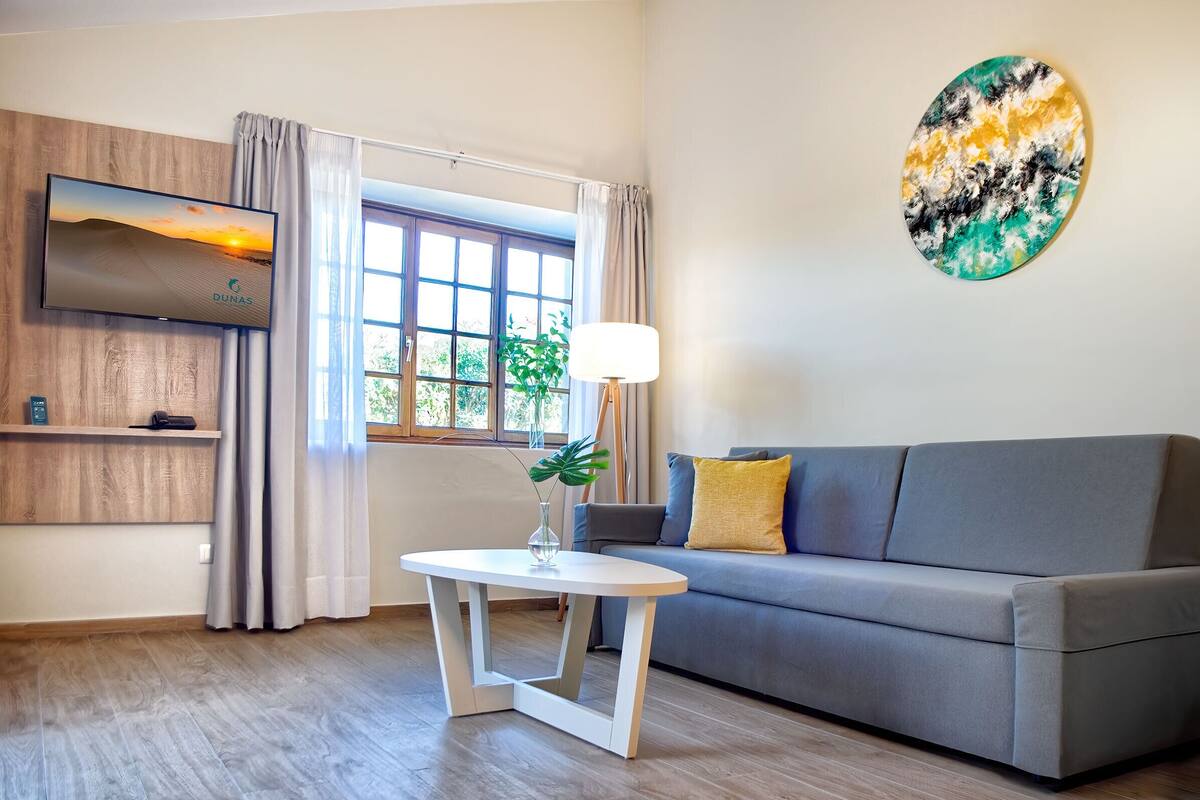 Ein modernes Wohnzimmer mit grauem Sofa, einem weißen Tisch, einem an einer Holzvertäfelung montierten Fernseher und einem Fenster mit Vorhängen.