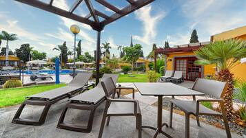 Un patio al aire libre con sillones y mesas, una piscina al fondo y cielo despejado.