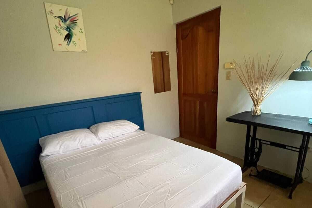 A bedroom with a bed, a desk, a chair, a door, and a wall-mounted lamp.