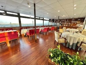 A restaurant with tables and chairs, a view of the water, and a large plant in the foreground. - Linjiang International Cruise Hotel (Shanghai)