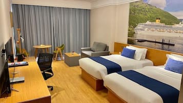 Habitación de hotel con dos camas, un escritorio con computadora, una silla, una mesita y un amplio ventanal con vista a un crucero.