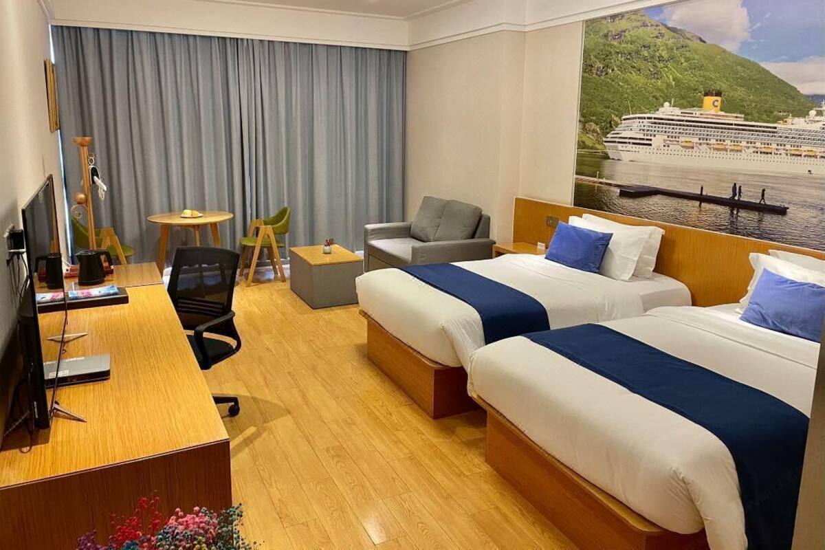 Habitación de hotel con dos camas, un escritorio con computadora, una silla, una mesita y un amplio ventanal con vista a un crucero.