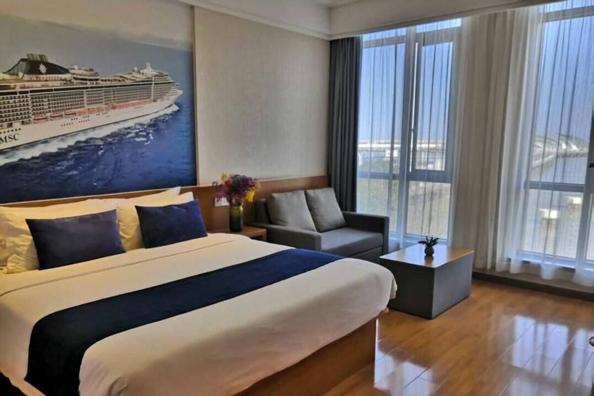 Una habitación de hotel con una cama grande, un sofá, una mesita y un cuadro de un crucero, además de vistas al exterior.