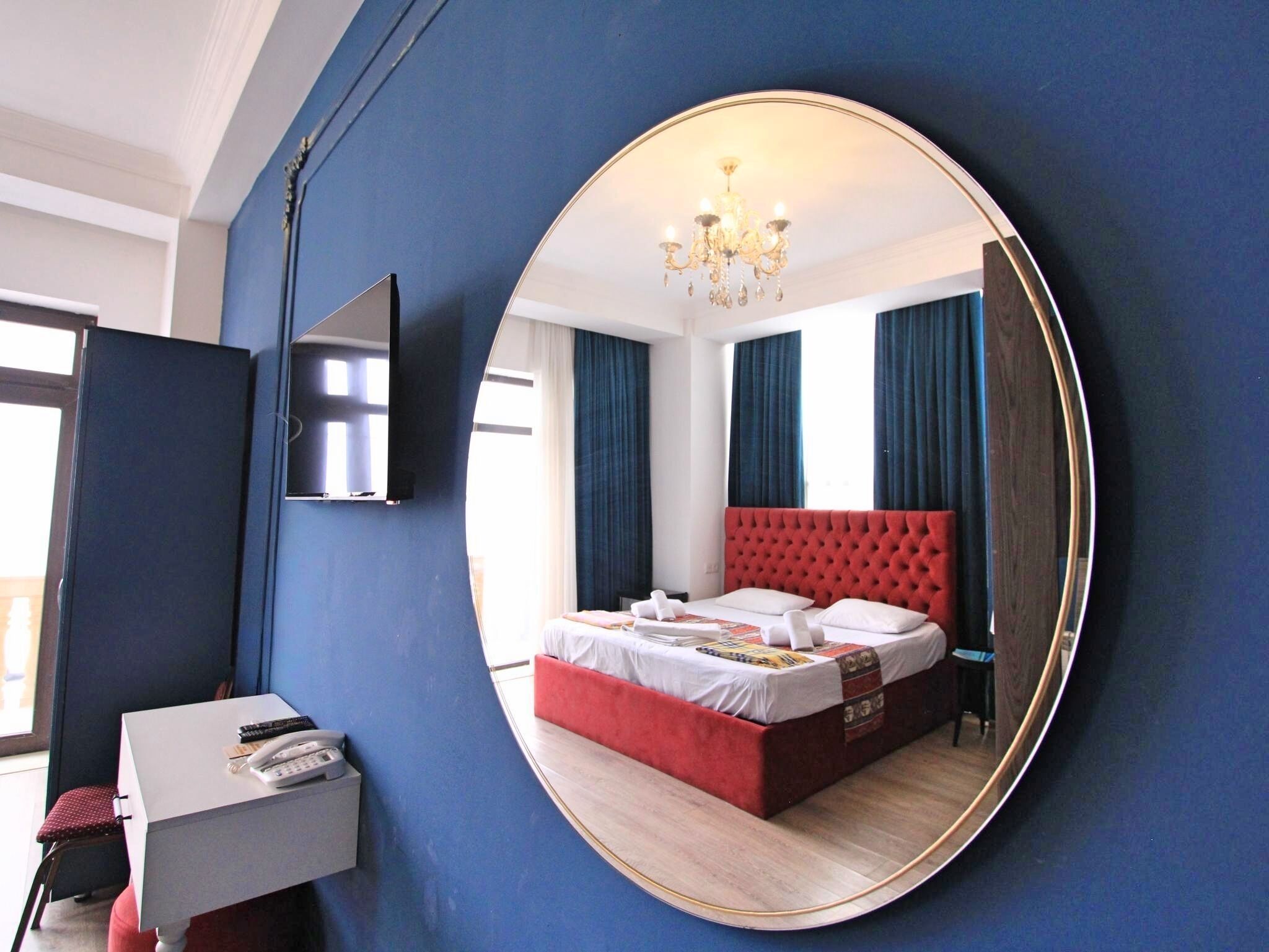 Een slaapkamer met een grote spiegel, een bed met een rode hoofdeinde, een bureau met een telefoon, en een tv aan de muur bevestigd.