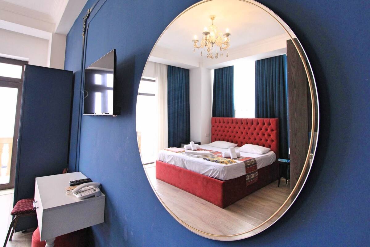 A bedroom with a large round mirror reflecting the room, a bed with a red headboard, a small desk with a telephone, and a TV mounted on the wall.