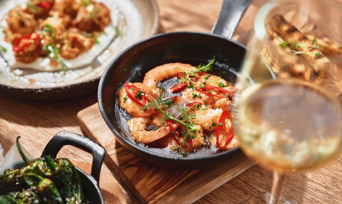 A pan with shrimp and vegetables, a glass of white wine, and a wooden table.