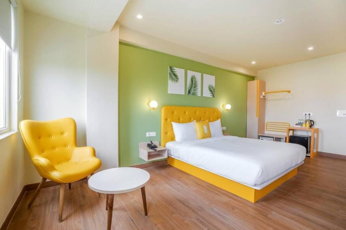 A bedroom with a bed, a yellow armchair, a small table, and a nightstand with a lamp.