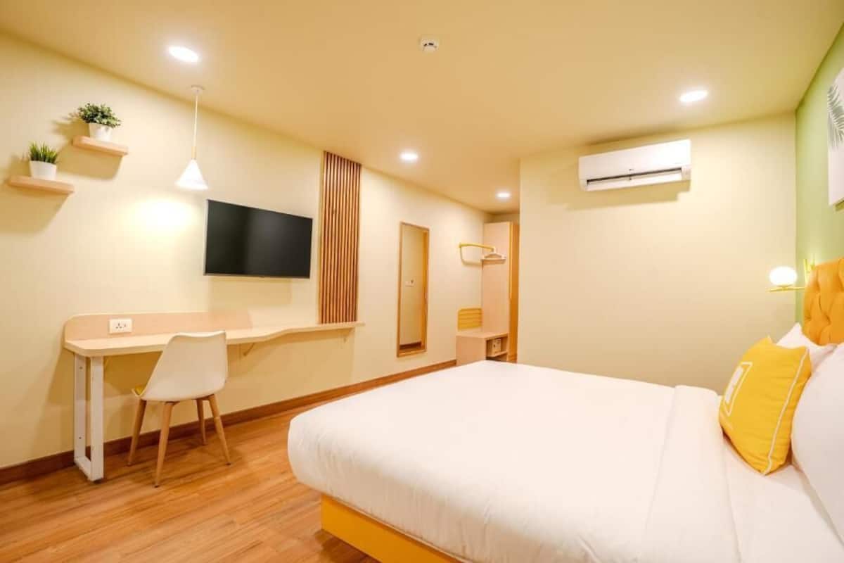 A hotel room with a bed, a desk, a chair, a TV, and an air conditioning unit.