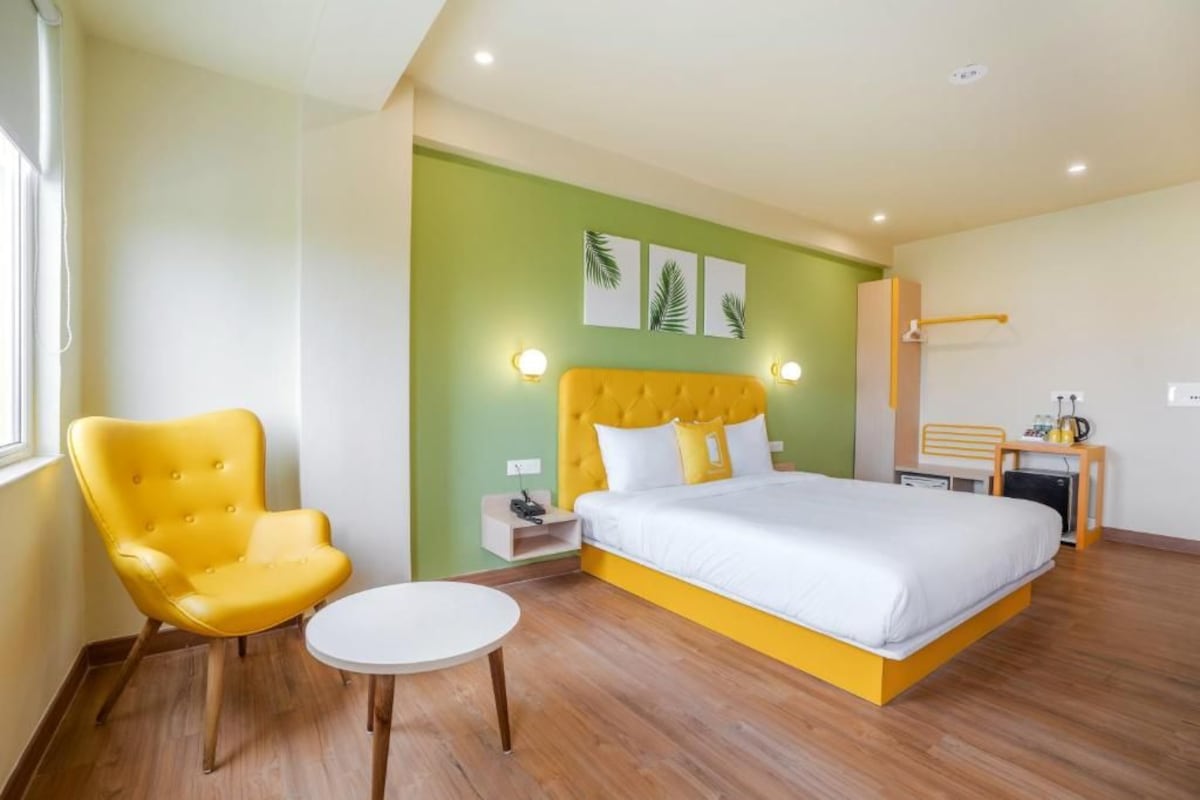 Un dormitorio con una cama, un sillón amarillo, una mesita y una mesita de noche con una lámpara.