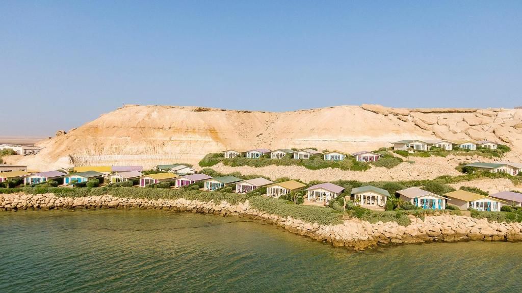 A coastal resort with villas along a rocky shoreline, clear skies, and a large cliff in the background.