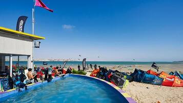 A pool area with a flag, a building with a flag, and a beach in the background.