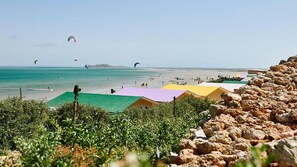 Una spiaggia con ombrelloni colorati, uno sfondo roccioso e aquiloni nel cielo.