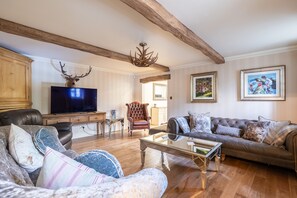 A living room with a television, leather sofas, a glass coffee table, wooden beams, and a chandelier.