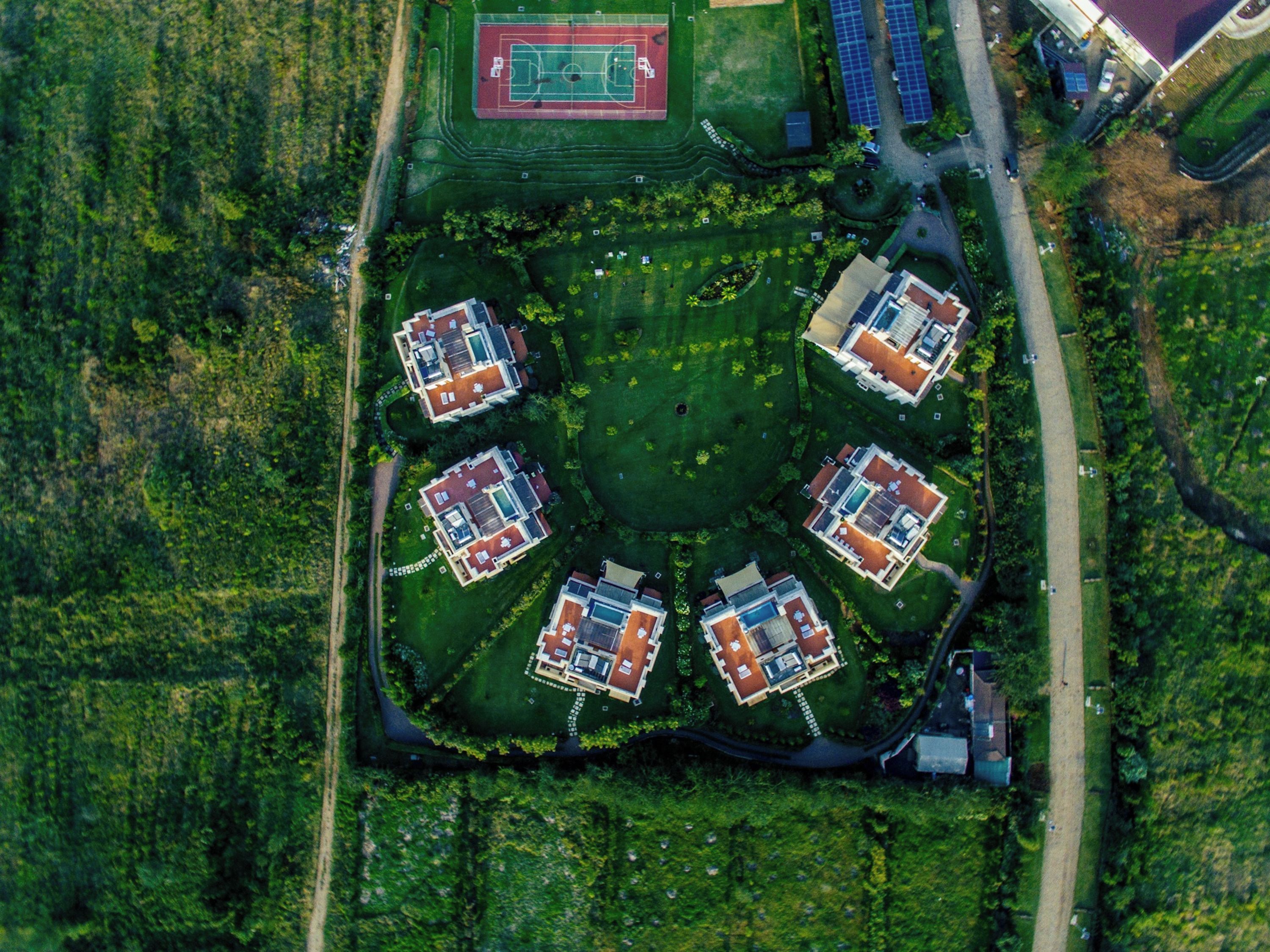 An aerial view of a residential area with six houses arranged in a circular pattern around a central green space, a sports field, and solar panels.