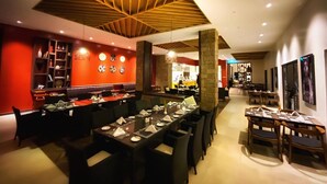 Un restaurante con un interior moderno, que presenta una zona de comedor central con mesas preparadas para el servicio, una pared de acento roja con platos decorativos y un diseño de techo de madera.