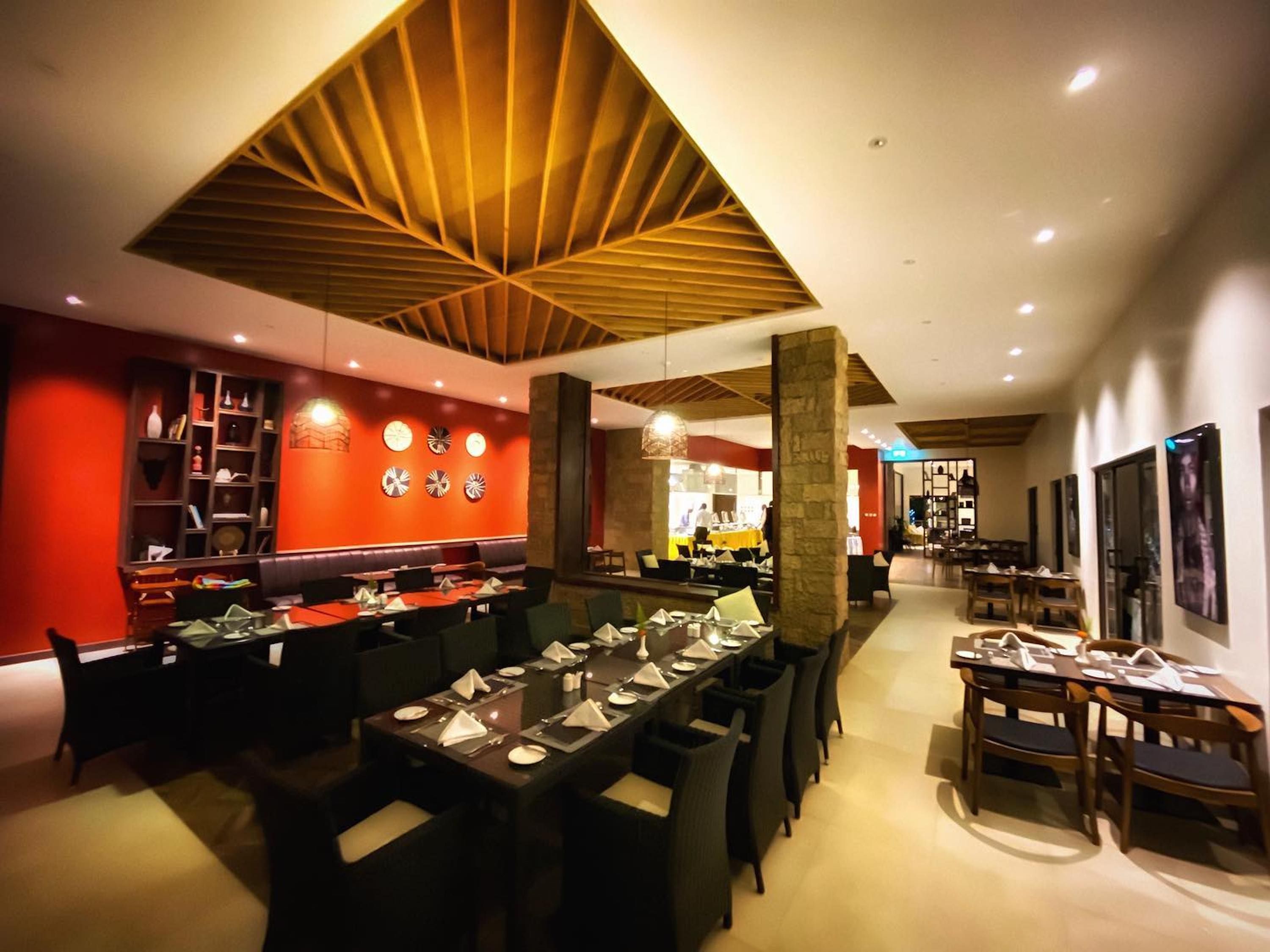 A restaurant with a modern interior, featuring a central dining area with tables set for service, a red accent wall with decorative plates, and a wooden ceiling design.