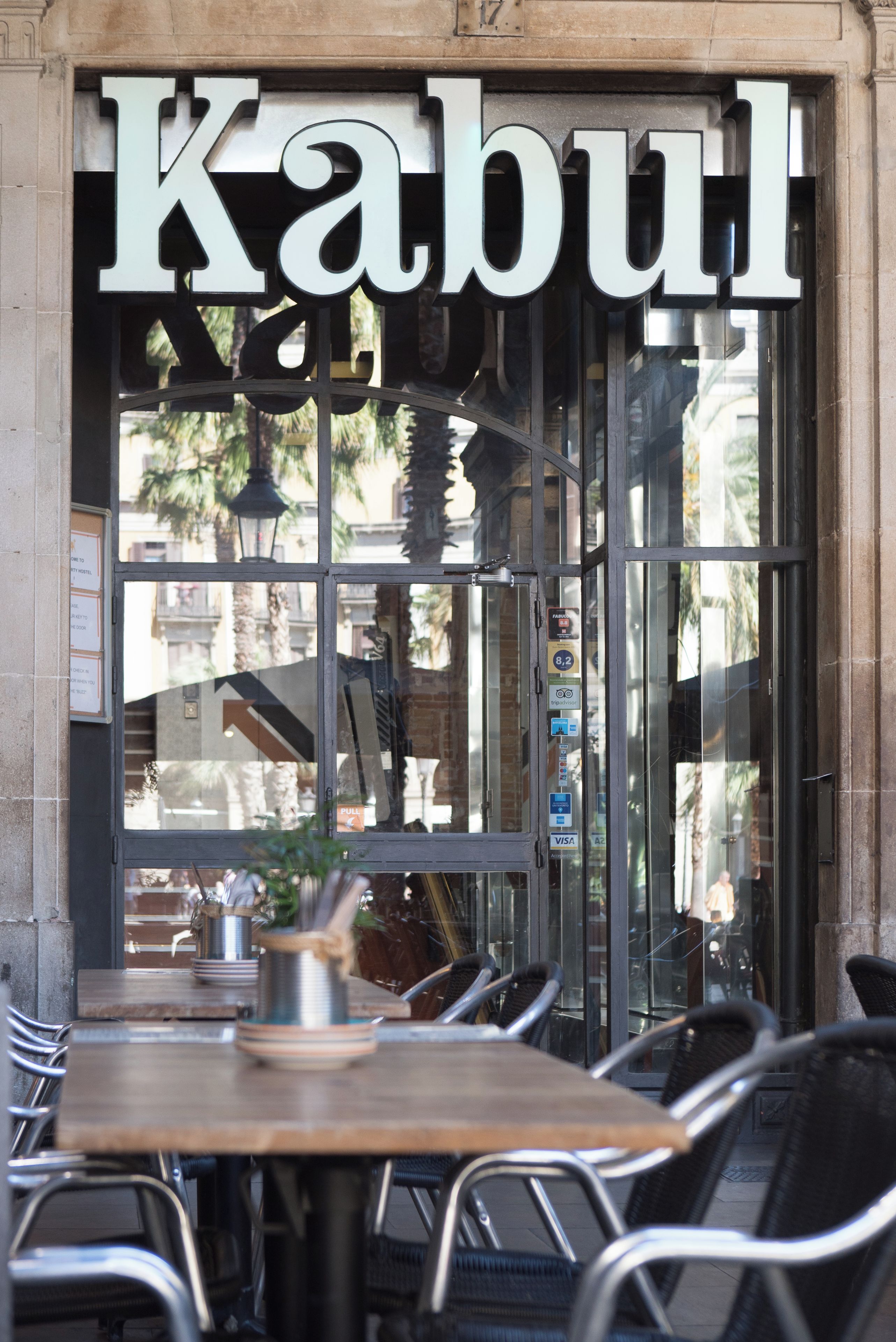 Entrada de restaurante com o nome 'Kabul' em letras grandes, porta de vidro e mesas externas com cadeiras de metal.
