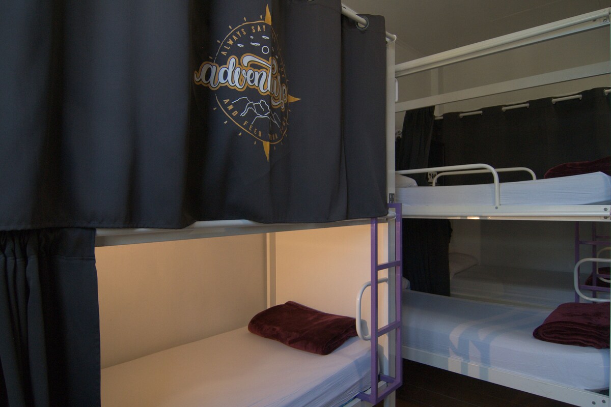 A bunk bed room with a curtain featuring a logo, a bed with a red blanket, and a white mattress.