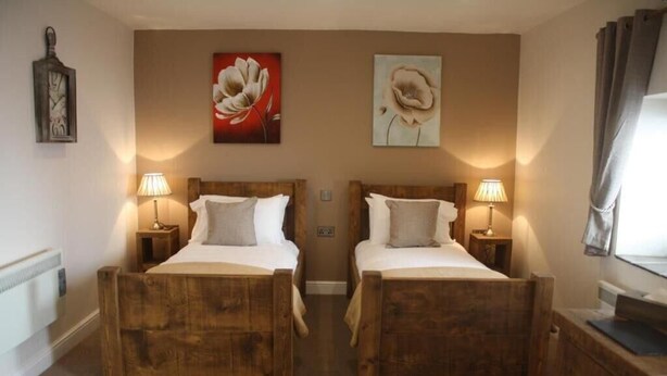 A double room with two wooden beds, each with a nightstand and lamp, and two large floral paintings on the wall. - Woodman Inn (Bedale)