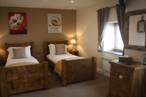 A bedroom with two wooden beds, a nightstand with a lamp, a dresser, and a window with curtains. - Woodman Inn (Bedale)