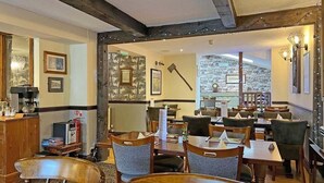 A restaurant with wooden tables and chairs, framed pictures on the walls, and a stone wall in the background. - Woodman Inn (Bedale)