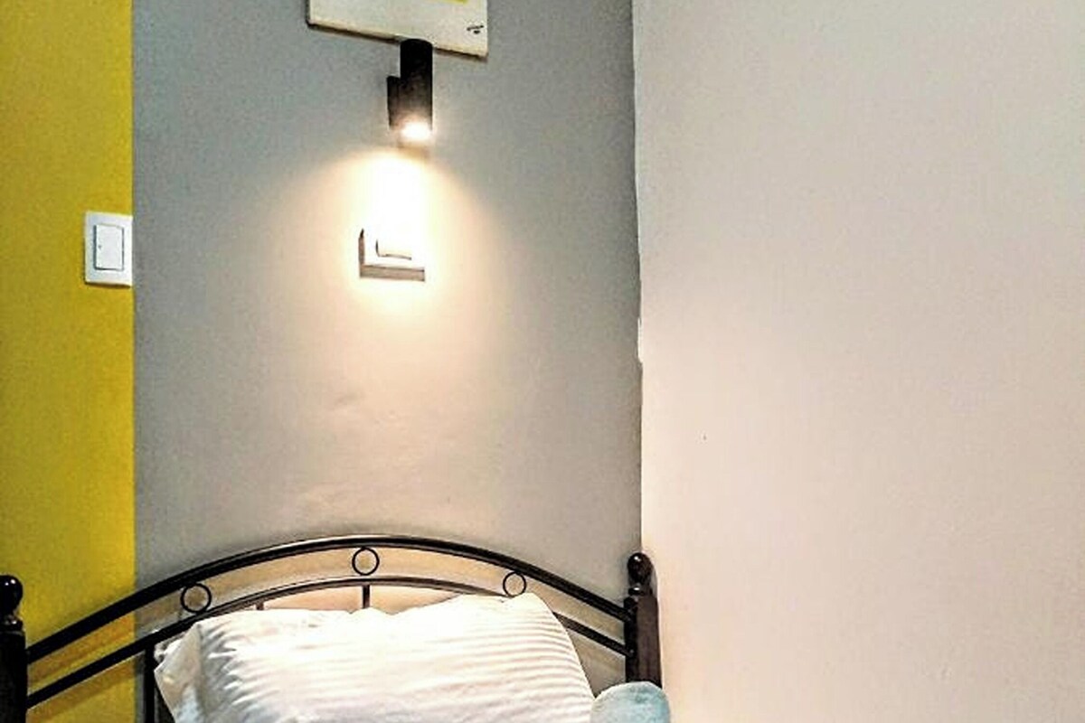 A neatly made bed with a pillow and rolled towels, a wall-mounted light, and a partial view of a yellow wall.