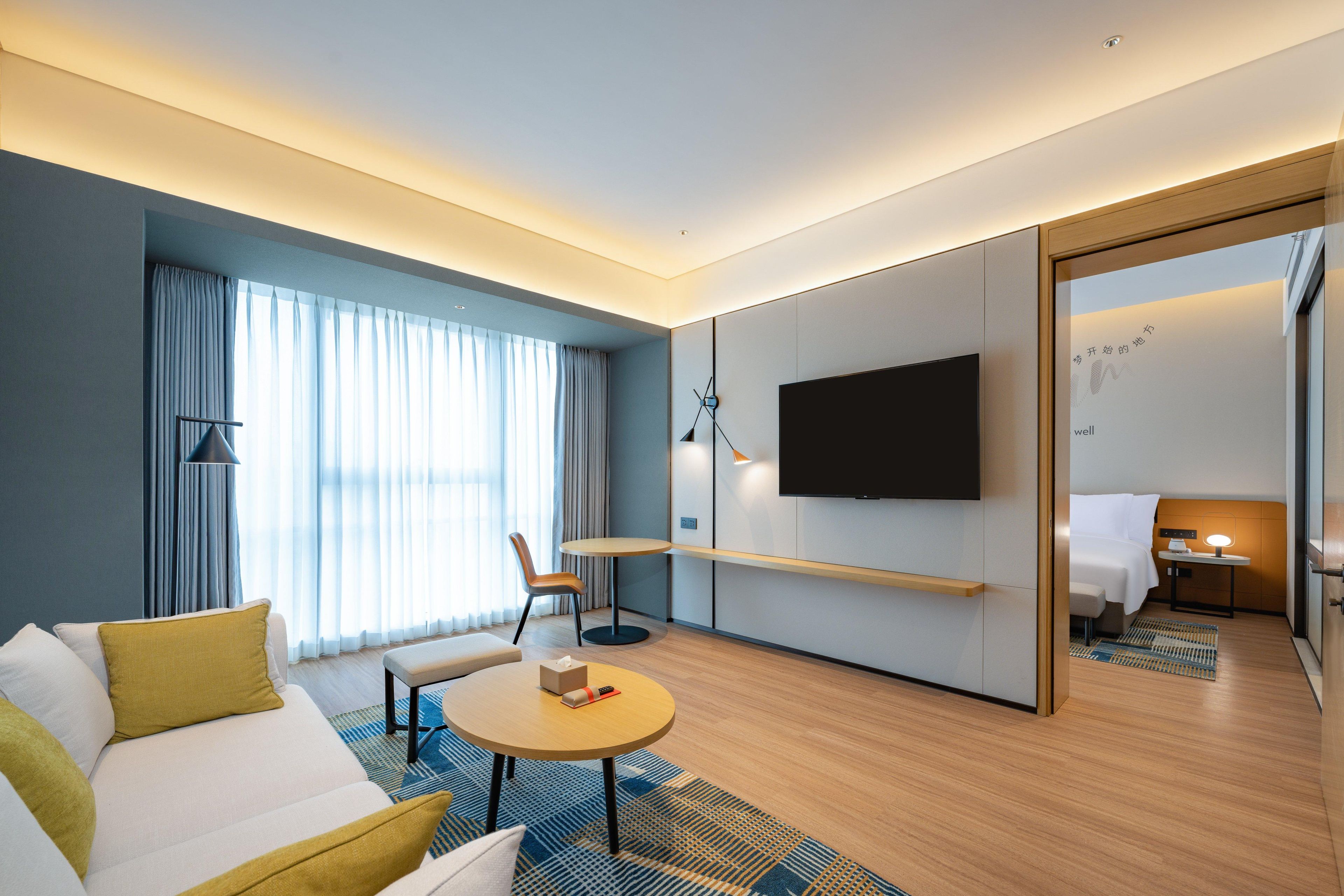 Una habitación de hotel moderna con sofá, una mesa redonda, televisión de pantalla plana, escritorio con lámpara y una cama.