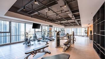 A modern gym with various exercise equipment, large windows, and a high ceiling.
