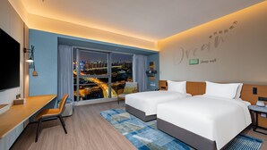 Habitación de hotel con dos camas, un escritorio, un televisor y un ventanal con vistas a la ciudad.