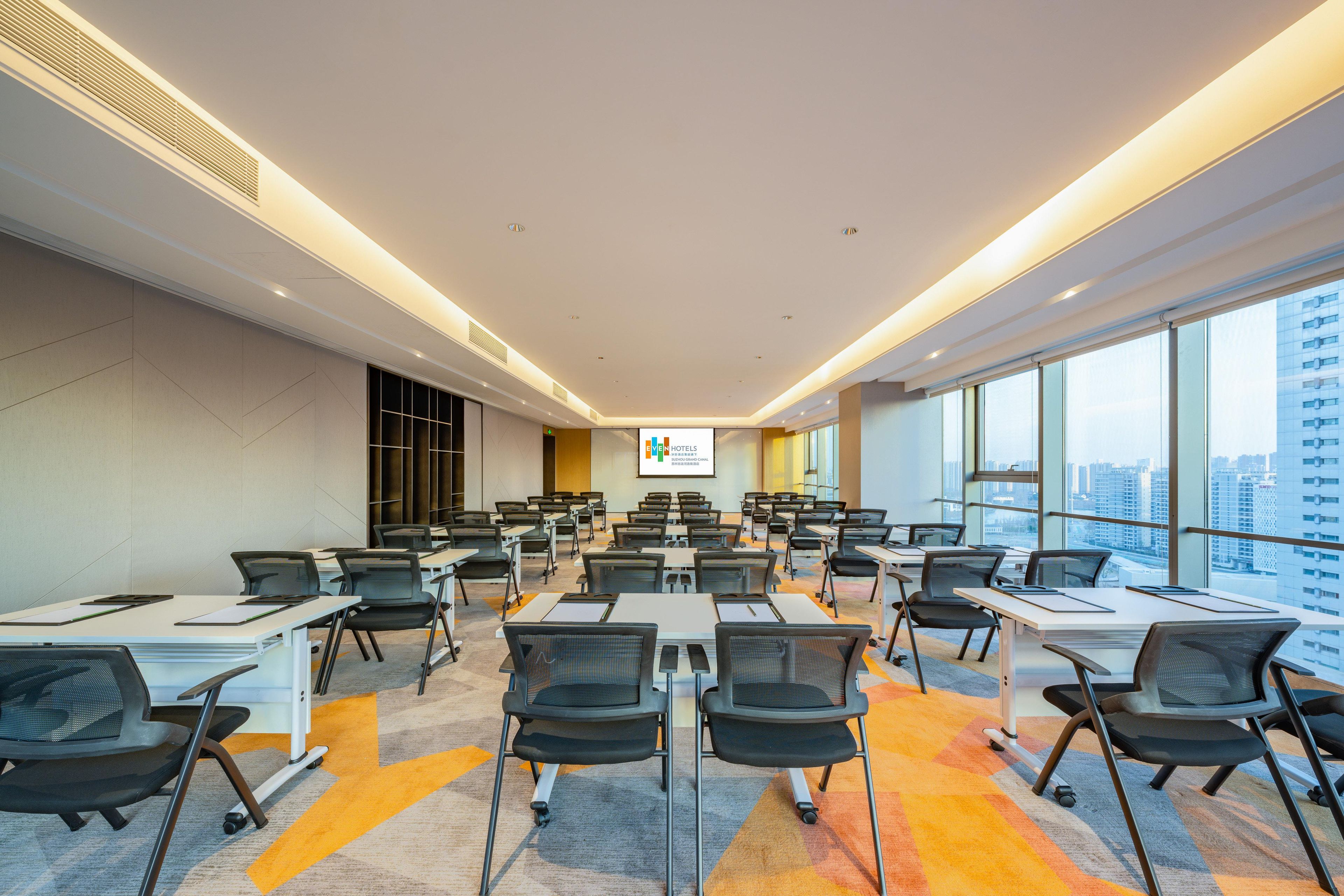 A conference room with rows of tables and chairs, large windows with a city view, and a presentation screen.