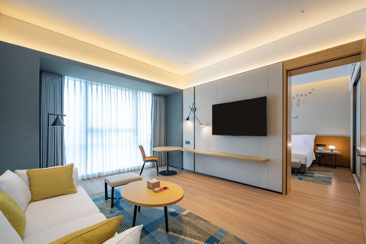 A modern hotel room with a sofa, a round coffee table, a flat-screen TV, a desk with a lamp, and a bed.